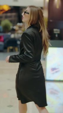 Uzun saçlı zarif bir kadın modern bir şehir merkezinde siyah deri bir elbise giyerek güvenle yürüyor. Modern Şehir Merkezi 'nde Siyah Deri Elbise Giyen Şık Bir Kadın. Dikey video.
