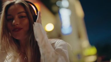 Genç bir kadın kulaklıkla müzik dinliyor, geceleri şehrin atmosferinin tadını çıkarıyor. Beyaz bir kıyafet giyiyor, gülümsüyor ve parlak ışıklı mağazaların önünde duruyor. Genç Kadın Müzikten Hoşlanıyor