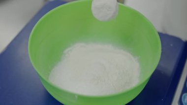 Şekerin yeşil bir kaseye döküldüğü yakın plan bir görüntü, zıt bir mavi yüzeye ayarlanmış, pişirme hazırlığının dinamik hareketini yakalıyor. Pişirmek için Yeşil Kase 'ye Şeker Dökmek