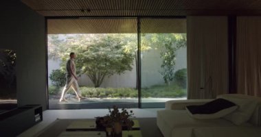 Huzurlu bir görüntü, huzurlu bir bahçede, modern bir oturma odasının yanında gezinen bir adamı gösteriyor. Doğal ışık ve bereketli yeşillik yalnızlığın sessiz anlarını vurgular..