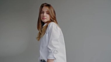 Beyaz gömlekli ve kot pantolonlu genç bir kadının portresi. Tarafsız bir arkaplan karşısında duruyor. Dengeli bir bakışla. Beyaz Gömlekli Genç Kadın Stüdyoda Gelişigüzel poz veriyor