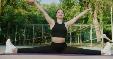 Spor elbiseli atletik bir kadın egzersiz minderinde bacağını ikiye ayırıyor, ellerini kaldırıyor ve parkta vücudunu esnetiyor. Genç bayan dışarıda egzersiz yapıyor ve esneklik eğitimi alıyor..