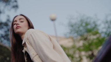 Rahat bir yüzü olan genç bir kadın güneşli bir günde bej bir ceketle dışarıda poz veriyor. Arka planda ağaçlar ve gökyüzü görülebilir. Rahat Genç Kadın Rastgele Kıyafetlerle Dışarda Poz Veriyor