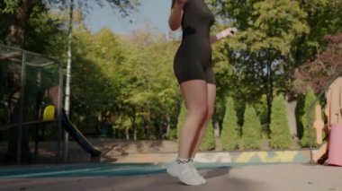 Sporcu kıyafeti giymiş fit bir genç kadın güneşli bir parkta ip atlayarak egzersiz yapıyor. Sağlık ve zindeliğin yaşam tarzını gösteriyor. Genç Kadın Atlama İpi 'nde güneşli bir günün tadını çıkarıyor.