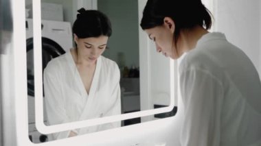Beyaz bornozlu genç bir kadın iyi aydınlatılmış banyo aynasına yansıyor ve sabah rutinine başlıyor. Sakin ve huzurlu bir an yakalandı. Banyo Aynasında Yansıyan Kadın, Sabah Rutini