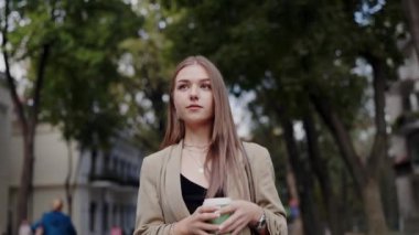 Şık takım elbiseli genç profesyonel bir kadın elinde kahve fincanı, şehir yaşam tarzını ve profesyonel güveni somutlaştıran bir şehir parkında yürüyor. Genç Kariyer Kadını Lush City Park 'ında Yürüyor