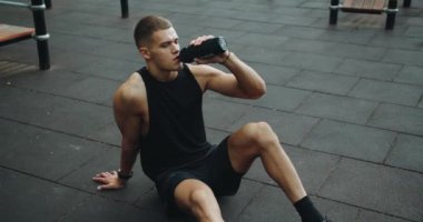 Sporcu adam antrenmana ara veriyor, yerde oturuyor, dinleniyor, susamış hissediyor ve spor sahasında şişeden su içiyor. Sağlıklı yaşam tarzı kavramı. Güç, motivasyon. Açık hava spor salonu.