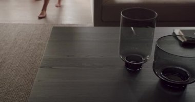 Koyu ahşap bir masanın üzerinde iki cam kasenin olduğu minimalist bir sahne, çıplak ayakların kısmi görüntüsü ve oturma odası arka planı, sakin, sade bir atmosfer taşır..