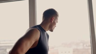 Formda, genç bir adam spor yaparken spor salonunun penceresinden dışarı bakıp fitness hedeflerini ve başarılarını yansıtıyor. Odaklanmış Erkek Sporcu Dinleniyor ve Spor Salonu Penceresine Bakıyor