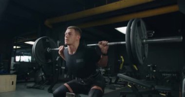 Genç bir erkek sporcu, iyi donanımlı bir fitness merkezinde spor ekipmanlarıyla çevrili ağır bir çömelme sahnesinde yoğun bir şekilde odaklanır. Modern Spor Salonu 'nda Ağır Çömelmeler Yapan Odaklı Sporcu