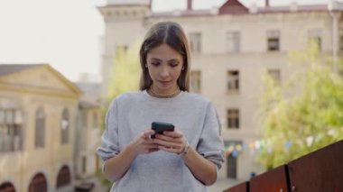 Genç bir kadın arka planda belediye binaları olan bir balkonda dikilirken akıllı telefonuna odaklanıyor. Şehir Caddesi Balkonunda Akıllı Telefon Kullanan Genç Kadın