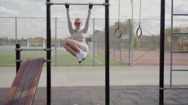 Spor giyim sektöründe formda genç bir kadın açık hava spor salonunda aktif bir yaşam tarzını gösteren yatay barlara asılıyor. Genç Kadın Spor Salonu Barlarında Egzersiz Yapıyor