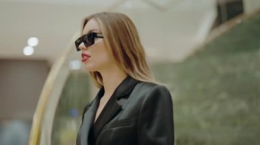 Siyah ceketli ve güneş gözlüklü zarif bir kadın, ev ortamında kendine güven ve tarz yayıyor. Modern bir çevrede yakalanmış, özgüvenli bir tavırla ileri moda bir görünüm sergiliyor..