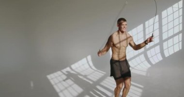 Atletik adam, parlak ışıklandırılmış bir stüdyoda atlama halatı egzersizi yapar, zindelik ve kararlılık gösterir. Sunlit Stüdyo Alanında Genç Atletik Atlama İpi