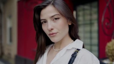 Şehir ortamında sakin ve kendinden emin genç bir kadının portresi. Yumuşak bir odak noktası ve şehir arkaplanı var. Şehir Çevresindeki Serene Genç Kadın Kendine Güvenle Bakıyor