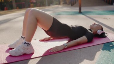 Güneşli bir günde parkta spor yaptıktan sonra dinlenen genç bir kadın. Genç bir kadın, parktaki pembe yoga minderinde dinleniyor, kulaklık ve spor kıyafetleri takıyor, vücut bulmuş spor kıyafetleri giyiyor ve rahatlıyor..