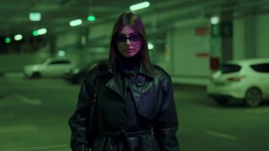 Güneş gözlüklü, uzun saçlı genç kadın model yeraltı otoparkında poz veriyor. Deri ceketli kadın park halindeki arabalara karşı büyüleyici bir görsel birliktelik yarattı. Şık kız..