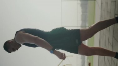 Spor giyimli bir erkek sporcu esniyor, koşuya hazırlanıyor. Odaklanmış ve elinden geleni yapmaya kararlı görünüyor. Koşu seansından önce esnemeye odaklanmış erkek sporcu. Dikey video.