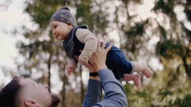 Neşeli genç bir baba, güler yüzlü bebeğini yemyeşil bir parkta havaya fırlatır. Sevgi, neşe ve baba-çocuk bağı gösterir. Neşeli Baba, parkta güneşli bir günde bebeğini kaldırıyor.