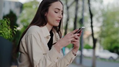 Parkta oturan genç bir kadın, akıllı telefonunu kullanıyor. Rahatlamış ve cihazıyla meşgul görünüyor, doğanın ortasında huzurlu bir anın tadını çıkarıyor. Akıllı Telefon Kullanan Genç Kadın