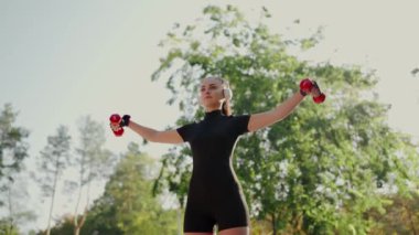Odaklanmış genç bayan sporcu, kollarını güçlendirmek için dambıllar kullanarak parkta egzersiz yapıyor. Dambıllarla parkta egzersiz yapan genç bir kadın.