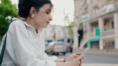 Şehrin kalabalık bir caddesinde otururken akıllı telefonunu kullanan bir kadının yan profili. Yaz günü şehir caddesinde mesajlaşan genç bir kadın. Cep Telefonu Kullanılıyor
