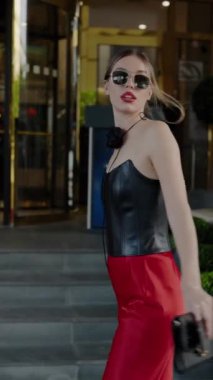 Kırmızı elbiseli ve güneş gözlüklü şık bir kadın, debriyaj tutuyor, lüks bir otelin önünde duruyor. Lüks Otelin dışında Kırmızı Elbiseli Zarif Kadın