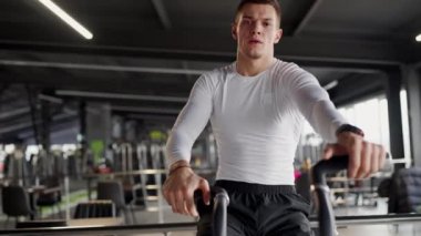 Dar beyaz gömlekli genç bir erkek sporcu, iyi donanımlı bir spor salonunda sabit egzersiz bisikleti sürmeye odaklanmış. Spor salonunda Beyazlı Genç Adam Egzersiz Bisikleti Kullanıyor