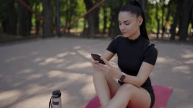 Açık hava fitness seansından sonra akıllı telefon kullanan genç bir kadın. Formda genç bir kadın parkta yoga minderinde oturur, akıllı telefonunu egzersizden sonra tutar. Su şişesi yan tarafta..