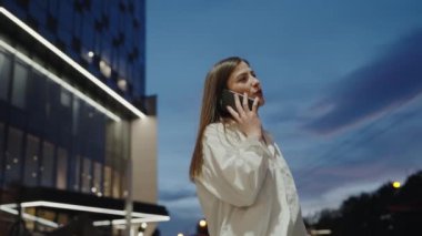Günbatımında açık havada bir telefon görüşmesi yapan sıradan giyinmiş genç bir kadın. Arka planda modern binalar var. Şehir Akşam Ayarlarında Akıllı Telefonla Konuşan Genç Kadın