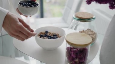 Modern, iyi ışıklandırılmış mutfak ortamında yaban mersini ve granola ile sağlıklı bir kahvaltı hazırlayan biri. Sahne sağlıklı beslenmeyi ve güne yeni bir başlangıç yapmayı teşvik ediyor..