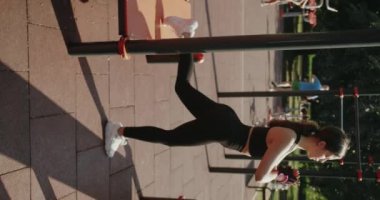 Genç bayan dışarıda egzersiz yapıyor ve esneklik eğitimi veriyor. Esnek bayan sporcu spor sahasında duruyor ve antrenmandan önce ısınırken metal korkuluklarda bacaklarını esnetiyor. Dikey video.