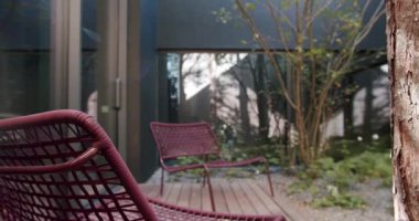 Konforlu bir kırmızı hasır sandalye, ağaç kabuğu dokusu ve çevresi yeşilliklerle çevrili çağdaş bir binanın manzarasına sahip modern açık hava verandası, huzurlu ve huzurlu bir atmosferi destekliyor..