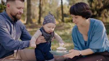 Üç kişilik genç bir aile parkta bir çocuğun doğum gününü kutluyor. Ebeveynler, bebeğin pastaya uzanmasını izler. Genç Aile Parkta Bebekle Doğum Günü Pikniği Yapıyor