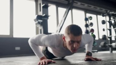 Genç bir erkek atlet iyi donanımlı bir spor salonunda yoğun bir dikkatle şınav çeker. Forma olan bağlılığı, kararlı ifadesi ve dinamik duruşunda belli. Şınav çeken genç adama odaklanmış.