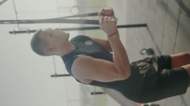 Spor salonunda direnç bantları kullanan odaklanmış genç bir erkek sporcu vücudunun üst kısmını güç antrenmanına bağlıyor. Jimnastik salonundaki genç adam direniş grubu egzersizleri yapıyor. Dikey video.