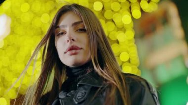 Işıkların arkasında kameraya poz veren güzel bir kadın. Kadın model şehir ışıklarıyla caddede dikilirken kameraya bakıyor. Gece lambalarının arka planında genç bir kadın var..