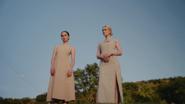 Açık havada açık mavi gökyüzüne karşı bej elbiseli iki zarif kadın. Görüntü, zarafet, tarz ve entelektüelliği doğal ışık altında resmediyor. Açık havada poz veren zarif kadınlar.