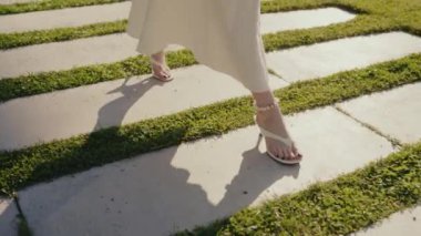 Dalgalı beyaz elbiseli, beyaz sandaletli, çimenler ve beton şeritlerle güneşli bir yolda yürüyen zarif ve zarif bir kadını yakından çek. Beyaz elbiseli zarif kadın yürüyor.