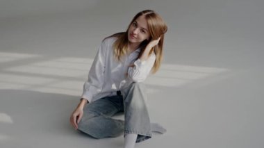 Beyaz gömlek ve kot pantolon giyen genç bir kadın, güneş ışığıyla yere oturur ve yumuşak gölgeler bırakır. 