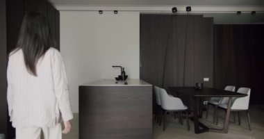 Bir kadın modern, minimalist bir mutfakta koyu ahşap dolaplar, temiz çizgiler ve şık mobilyalarla duruyor. Uzay karmaşıklık ve çağdaş tasarım yayar. Kadınla Minimalist Yemek Odası