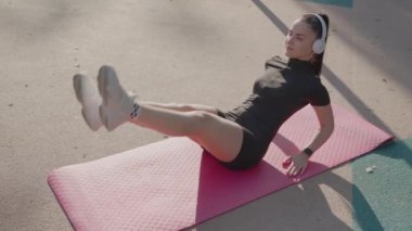 Spor kıyafetleri ve kulaklıklar takan odaklanmış genç bir kadın açık havada pembe bir paspasın üzerinde mekik çekiyor ve fitness gösterisi yapıyor. Genç Kadın Pembe Mat Üzerinde Çıtırtı Çalıyor