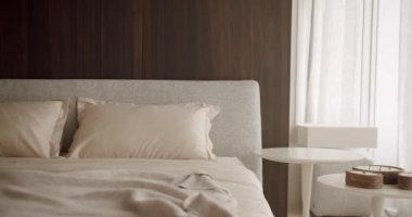 Huzurlu ve modern minimalist bir yatak odası, bej yastıklı rahat bir yatak ve ahşap bir yatak başlığı. Oda, sakin bir atmosfer için beyaz masalar ve yumuşak ışıklandırmalarla dolu..