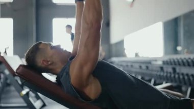 Atletik adam, modern spor salonundaki eğimli bir bankta ağır dambılları kaldırıyor. Vücut kaslarına odaklanmak için güç antrenmanı yapıyorlar. Spor salonunda dambılları kaldırmaya odaklanmış bir adam.