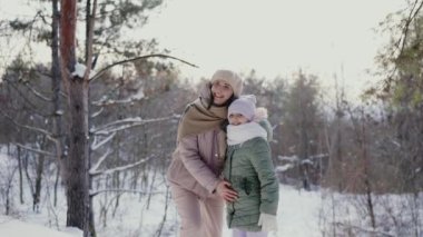 Neşeli genç bir anne, küçük kızını karlı bir ormanda kucaklıyor. Hem gülümsüyor hem de kışlık kıyafetlerle sarılıyor. Mutlu Anne ve Kız Karlı Orman 'da Kış Gününün tadını çıkarıyor