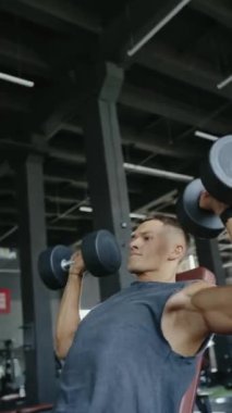 Spor salonunda dambılları kaldırmaya odaklanmış genç bir adam, dayanıklılık ve zindelik gösteriyor. Sahne kararlılığı, egzersizi ve sağlık ve refahı vurguluyor. Dikey