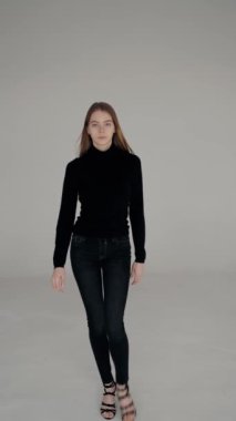 Genç bir kadın, siyah balıkçı yaka ve kot pantolon giymiş bir stüdyo ortamında kameraya doğru güvenle yürüyor. Stüdyoda yürüyen kendine güvenen genç kadın, sıradan moda. Dikey video.