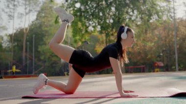 Atletik bir kadın, bir parkta kulaklıkla yoga minderinin üzerine uzanır. Arka planda ağaçlarla güneşli bir gün egzersizi. Açık havada egzersiz yapan, güneşli bir günde kulaklık takan genç bir kadın.