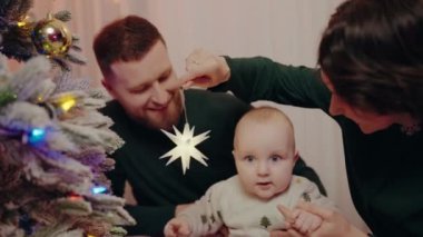 Sevgi dolu bir aile, Noel ağacını elinde yıldız süsü tutan bir bebekle süslüyor. Bu festival sahnesi, tatil sezonunun sıcaklığını, neşesini ve birlikteliğini yansıtıyor. Aile Noel Ağacı Dekorasyonu