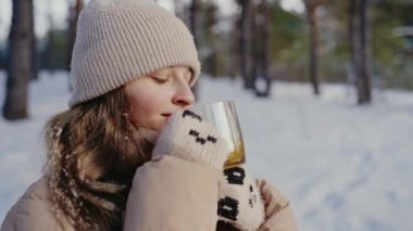 Örgü şapkalı genç bir kadın, sakin ve karlı bir ormanla çevrili ılık bir içeceği yudumluyor. Görüntü, huzur ve kış cazibesiyle doludur. Karlı Ormanda Sıcak Bir İçkinin Tadını Çıkaran Kadın
